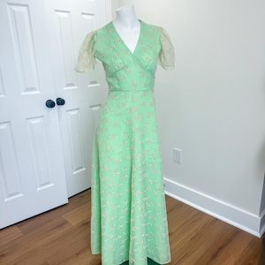 May include: A mint green vintage dress with a V-neckline and short, sheer sleeves. The dress features a delicate floral pattern in a lighter shade of pink. The dress is displayed on a mannequin.