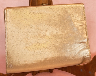 May include: A gold metallic clutch purse with a subtle shimmer. The purse has a simple, rectangular shape and a smooth, textured surface.