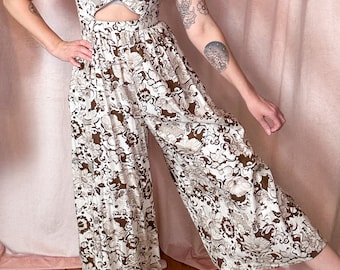 May include: A white and brown floral print jumpsuit with a cutout at the waist. The jumpsuit has wide legs and a sleeveless bodice.