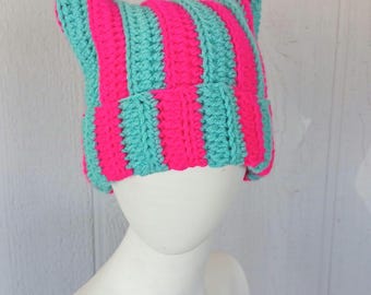 May include: A handmade, striped cat ear beanie in bright pink and teal. The crochet hat features vertical stripes and pointed ears, perfect for a playful look. The hat is displayed on a white mannequin head.