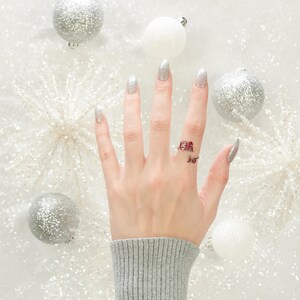 May include: A hand with white nail polish is wearing a silver ring with red letters that spell "Oh, the places you'll go!" The hand is surrounded by silver and white ornaments.