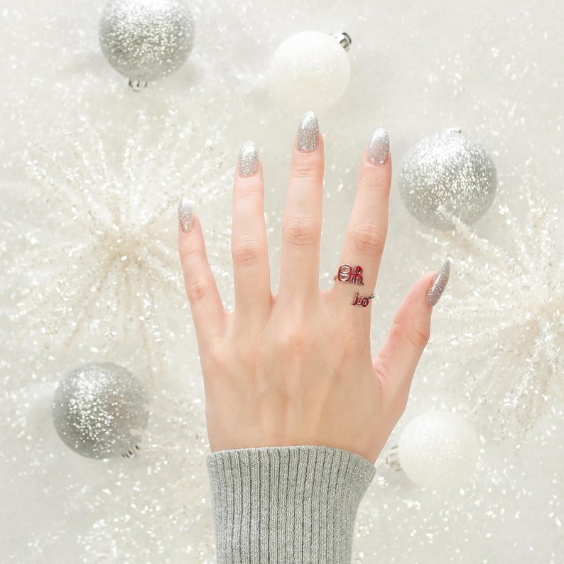 May include: A hand with white nail polish is wearing a silver ring with red letters that spell "Oh, the places you'll go!" The hand is surrounded by silver and white ornaments.
