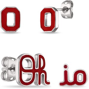 May include: Silver-toned stud earrings with red enamel accents. The earrings feature the letter "O" and the letters "OR" in a stylized font. The earrings are designed for pierced ears and are presented on a white background.