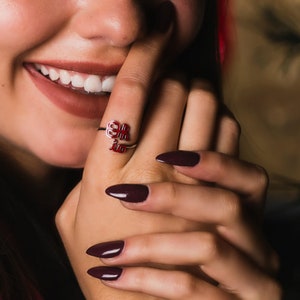 May include: A close-up of a person's hand wearing a silver ring with red Chinese characters. The ring is on the index finger. The person's nails are painted a dark burgundy color.