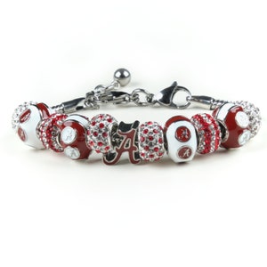 Puede incluir: Un brazalete de plata con abalorios rojos y blancos y un abalorio rojo y blanco con la letra 'A' en letra cursiva. Los abalorios est&aacute;n decorados con pedrer&iacute;a transparente.