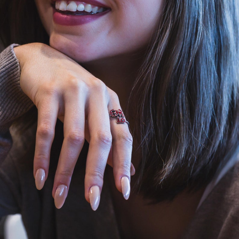 May include: A silver ring with red enamel letters spelling "Girl" on the band. The ring is worn on the index finger of a hand with light pink nail polish.