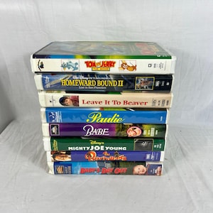 May include: A stack of VHS tapes featuring various family movies. Titles include "Tom & Jerry: The Movie", "Homeward Bound II", "Leave It To Beaver", "Paulie", "Babe", "Mighty Joe Young", "The Rescuers", and "Baby's Day Out". The tapes are in their original packaging.