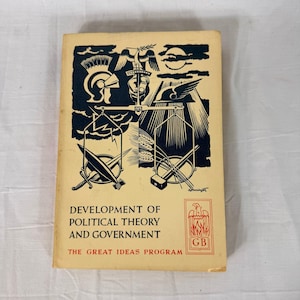 May include: A vintage book titled "Development of Political Theory and Government" with a cream-colored cover. The cover features a black and white illustration of a helmet, scales, swords, and other symbols. The text "The Great Ideas Program" is also visible.