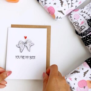 Puede incluir: Imagen con tem&aacute;tica de Halloween con una tarjeta de felicitaci&oacute;n con dos figuras de fantasmas y el texto "YOU'RE MY BOO!" rodeada de regalos envueltos. Los regalos est&aacute;n envueltos en papel blanco con calabazas rosas, murci&eacute;lagos y estrellas.