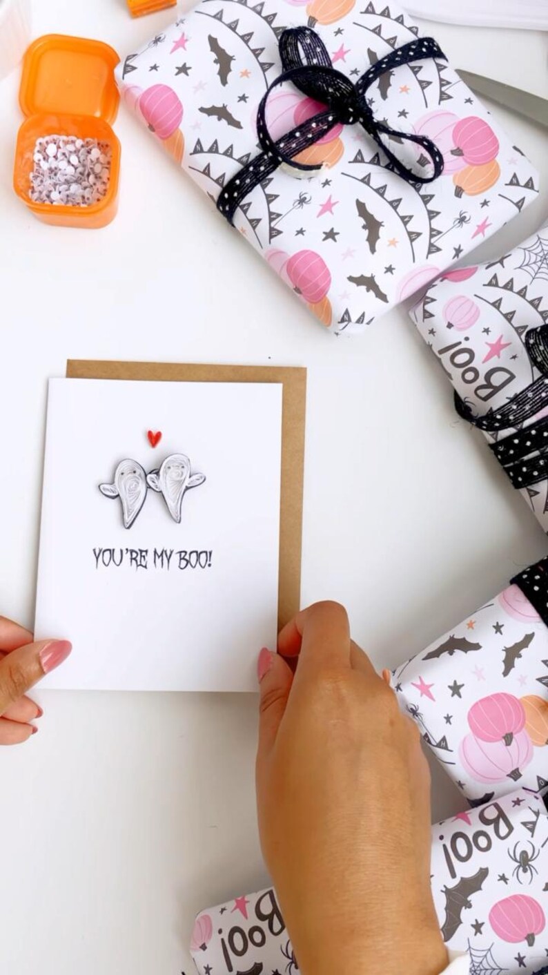 Puede incluir: Imagen con tem&aacute;tica de Halloween con una tarjeta de felicitaci&oacute;n con dos figuras de fantasmas y el texto "YOU'RE MY BOO!" rodeada de regalos envueltos. Los regalos est&aacute;n envueltos en papel blanco con calabazas rosas, murci&eacute;lagos y estrellas.