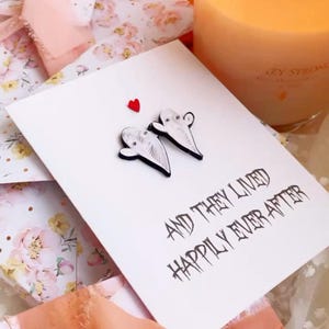 Puede incluir: Tarjetas de felicitaci&oacute;n blancas con adornos en forma de fantasma y el texto "AND THEY LIVED HAPPILY EVER AFTER". Tambi&eacute;n se ven una vela encendida y una cinta rosa. Las tarjetas est&aacute;n sobre una superficie con estampado floral.