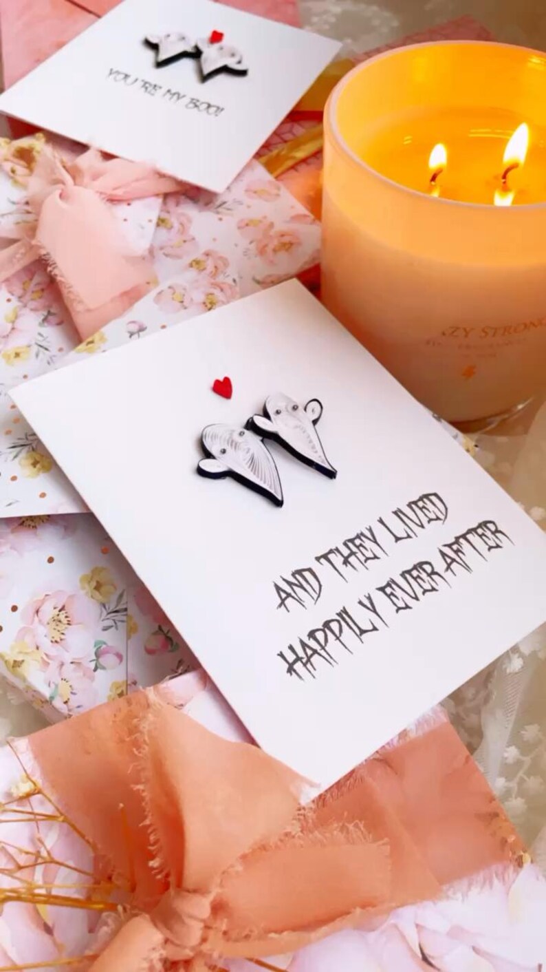 Puede incluir: Tarjetas de felicitaci&oacute;n blancas con adornos en forma de fantasma y el texto "AND THEY LIVED HAPPILY EVER AFTER". Tambi&eacute;n se ven una vela encendida y una cinta rosa. Las tarjetas est&aacute;n sobre una superficie con estampado floral.