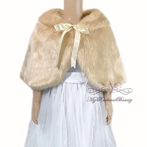 May include: A beige faux fur cape with a satin ribbon tied in a bow at the neck. The cape is worn over a white tulle dress.