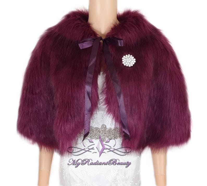 May include: A burgundy faux fur shrug with a satin ribbon tie and a jeweled brooch. The shrug is short and has a collar.