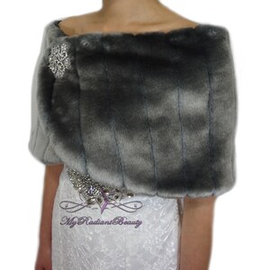 May include: Gray faux fur shrug with a silver rhinestone brooch. The shrug is draped over the shoulders of a model wearing a white lace dress.