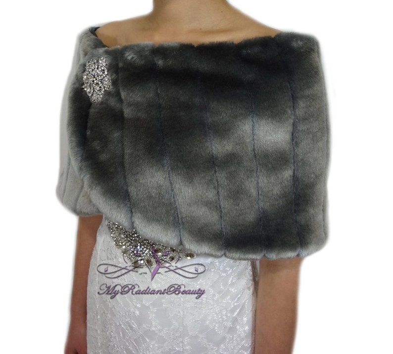 May include: Gray faux fur shrug with a silver rhinestone brooch. The shrug is draped over the shoulders of a model wearing a white lace dress.