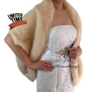 May include: A cream-colored faux fur shawl with a wide, fluffy collar. The shawl is draped over a white strapless dress with a lace pattern. A rhinestone belt is worn around the waist.