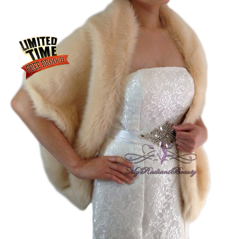 May include: A cream-colored faux fur shawl with a wide, fluffy collar. The shawl is draped over a white strapless dress with a lace pattern. A rhinestone belt is worn around the waist.