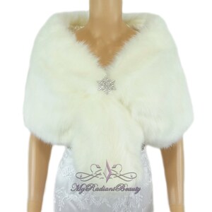 Puede incluir: Chal de piel sint&eacute;tica blanca con un broche de cristal en forma de copo de nieve. El chal est&aacute; drapeado sobre un maniqu&iacute; que lleva un vestido blanco.