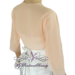 May include: Peach colored sheer chiffon bolero with long sleeves and a tie at the back. The bolero is shown on a white dress with lace detailing.