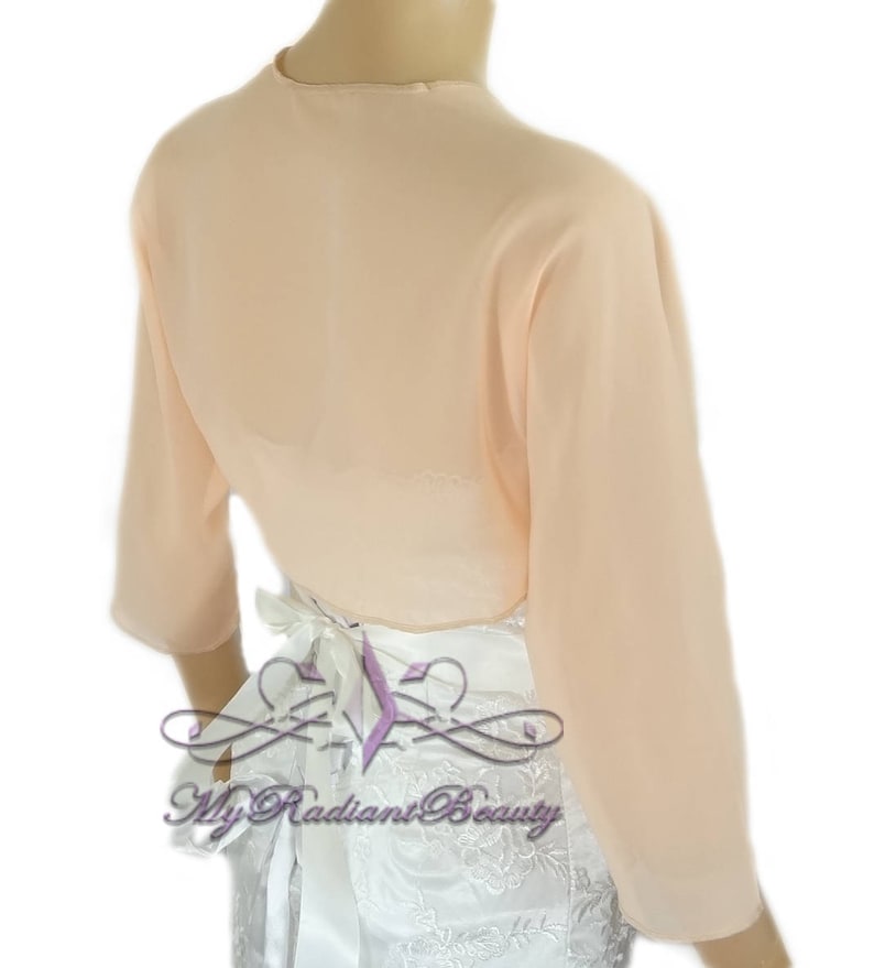 May include: Peach colored sheer chiffon bolero with long sleeves and a tie at the back. The bolero is shown on a white dress with lace detailing.