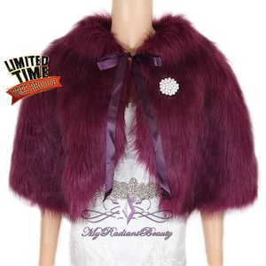 May include: A burgundy faux fur shrug with a satin ribbon tie and a rhinestone brooch. The shrug has short sleeves and a collar.