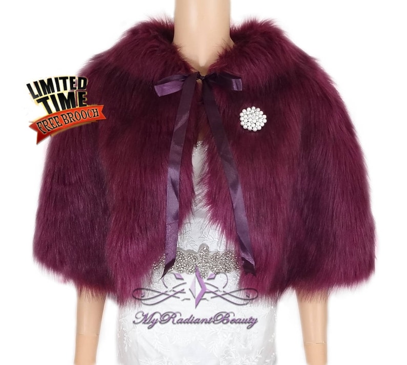 May include: A burgundy faux fur shrug with a satin ribbon tie and a rhinestone brooch. The shrug has short sleeves and a collar.