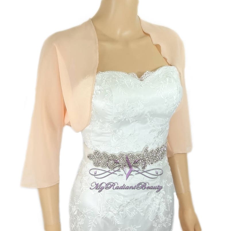 May include: Peach colored chiffon shrug with 3/4 sleeves. The shrug is draped over a white lace dress with a rhinestone belt.