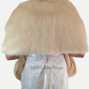 May include: A cream-colored faux fur shawl with a satin ribbon tie. The shawl is draped over a white dress with lace detailing.