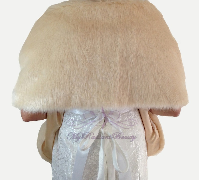 May include: A cream-colored faux fur shawl with a satin ribbon tie. The shawl is draped over a white dress with lace detailing.