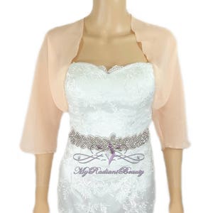 May include: Peach colored chiffon shrug with scalloped edges. The shrug is three-quarter length and has a relaxed fit.  The model is wearing the shrug over a white lace dress with a rhinestone belt.