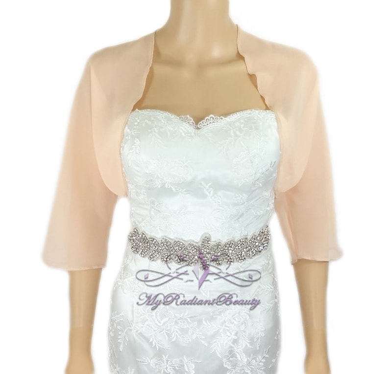 May include: Peach colored chiffon shrug with scalloped edges. The shrug is three-quarter length and has a relaxed fit.  The model is wearing the shrug over a white lace dress with a rhinestone belt.