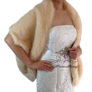 May include: A cream-colored faux fur shawl with a long, flowing design. The shawl is draped over a white strapless dress with a lace pattern. The dress has a white satin belt with a jeweled buckle.
