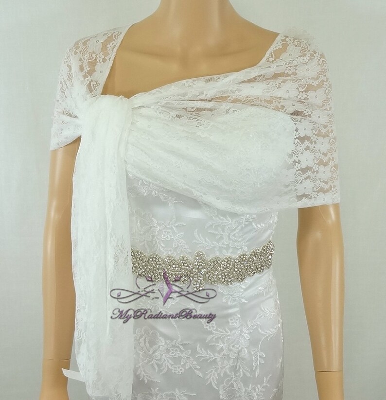 May include: White lace bridal shawl with a delicate floral pattern. The shawl is draped over a mannequin's shoulders and tied at the neck. The shawl is made of soft, sheer lace and would be a beautiful addition to any bridal ensemble.