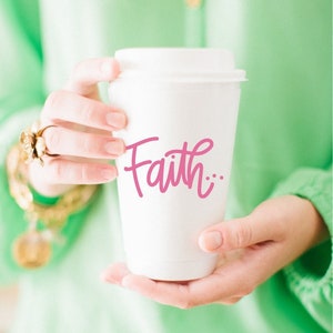 May include: A white to-go coffee cup with a pink cursive inscription that reads "Faith..." The cup is being held by a person wearing a light green shirt.