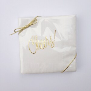 May include: White paper napkins with gold foil lettering that says "Cheers!" The napkins are wrapped with a gold ribbon and tied with a bow.