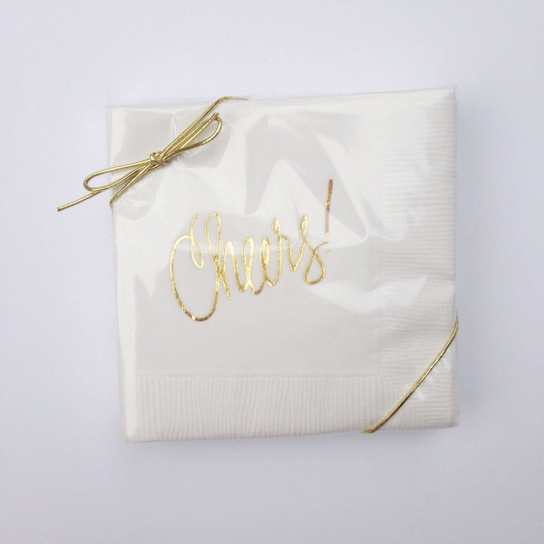 May include: White paper napkins with gold foil lettering that says "Cheers!" The napkins are wrapped with a gold ribbon and tied with a bow.