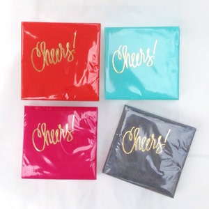 May include: Four square coasters with the word "Cheers!" written in gold lettering. The coasters are red, turquoise, pink, and gray.