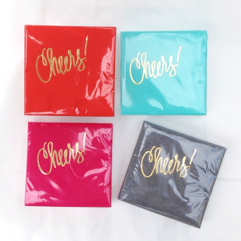May include: Four square coasters with the word "Cheers!" written in gold lettering. The coasters are red, turquoise, pink, and gray.