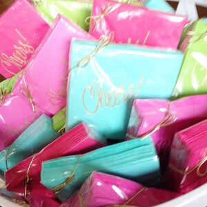 May include: A pile of pink, turquoise, and green paper napkins with gold lettering. The napkins are wrapped in clear plastic and tied with gold ribbon. The text on the napkins reads "Cheers!" and "Congrats!"