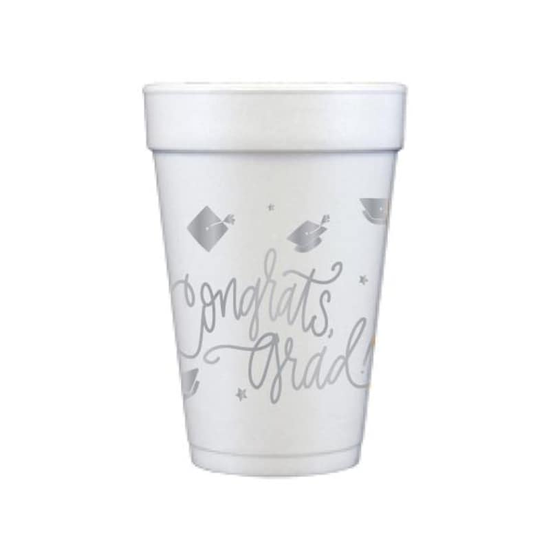 Puede incluir: Un vaso de espuma blanco con texto plateado y gr&aacute;ficos de gorros de graduaci&oacute;n. El vaso presenta las palabras "Congrats Grad" en una fuente de escritura, junto con peque&ntilde;as ilustraciones de estrellas y birretes. Ideal para celebraciones de graduaci&oacute;n.