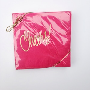 May include: Pink paper napkins with a gold "Cheers!" inscription, wrapped in clear plastic with a gold ribbon.