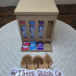 May include: A beige 3D-printed dispenser with four vertical slots containing Hawaiian Punch drink mix packets. The bottom tray holds three individual packets in red, blue, and purple. A wooden sign features three tree silhouettes and the text @3TreesStitchCo.