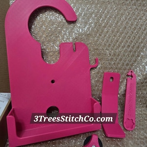 May include: A set of bright pink 3D-printed accessories, including a hanger-like structure, a rectangular piece, a small tool, and a triangular knob. The text "3TreesStitchCo.com" is printed on the main piece and the small tool.