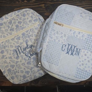 May include: Two quilted bags with floral and patchwork patterns. One bag features a blue floral design with "Marilyn Shaw" embroidered in blue. The other bag has a patchwork design with the initials "CWN" embroidered in blue. Both bags have a zipper closure.