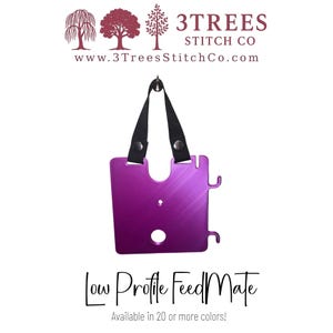 May include: A purple metal Low Profile FeedMate tool featuring a square shape with rounded corners, a central hole, and a smaller hole above it. It includes a black fabric strap attached with two silver metal snaps and two side hooks.