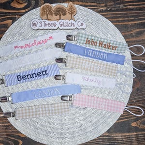 May include: A collection of personalized fabric pacifier clips in various colors and patterns, including gingham and solid pastels. Each clip features embroidered names such as meri claire, John Walker, Landon, Bennett, James, Rhenley, and Tatum, with metal clasps and white loops.