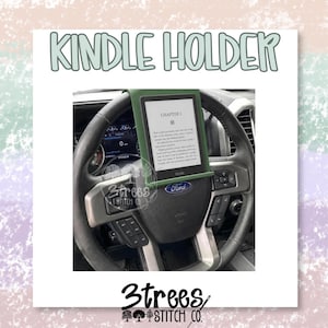 May include: A sage green Kindle holder mounted onto a black Ford steering wheel. The holder features a rectangular frame that secures an e-reader displaying text titled Chapter 1. The text KINDLE HOLDER appears in large letters above the device.