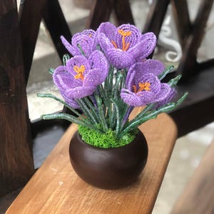 May include: A handcrafted beaded flower arrangement featuring purple crocus blooms with orange centers, green stems, and a brown ceramic pot. The arrangement sits on a wooden surface, showcasing the intricate beadwork and vibrant colors.