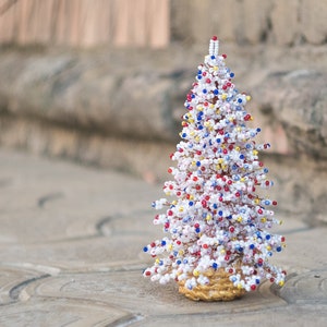 Peut inclure: Une sculpture d'arbre décorative fabriquée à partir de fil de cuivre torsadé orné de nombreuses petites perles blanches, rouges et bleues. La structure conique repose sur une base circulaire dorée texturée, créant un effet perlé dense.